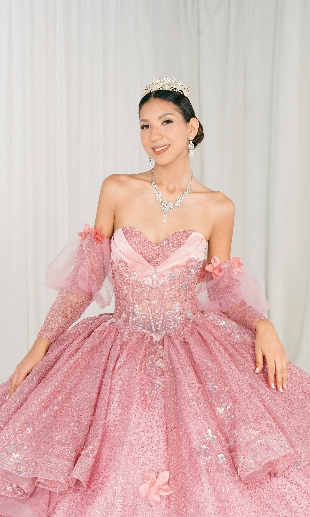 A young woman wearing the Quinceanera Dress 8385J By Cinderella Couture, featuring a sparkling pink corset bodice, with a tiara and jewelry, poses in front of a light-colored curtain background.